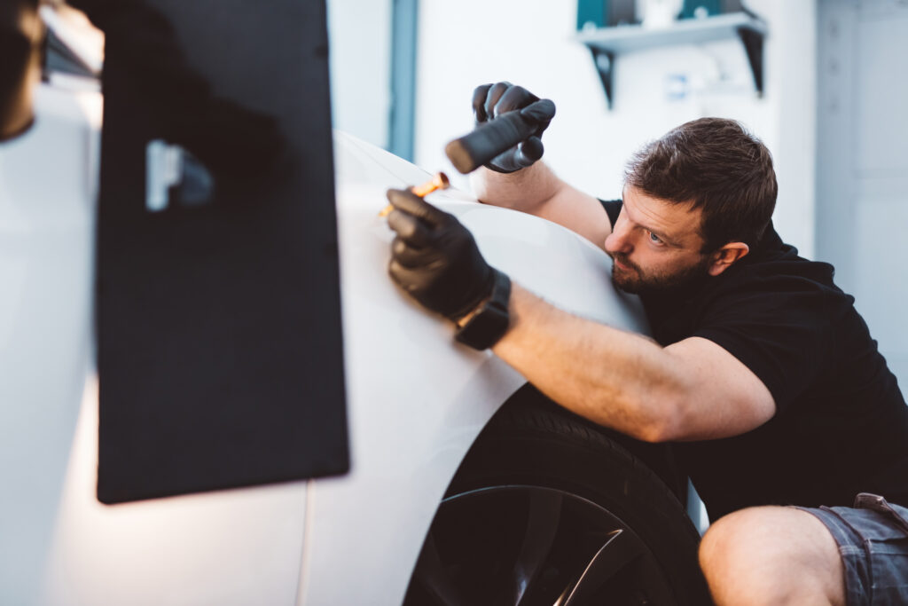A technician performing paintless dent removal on a car panel using specialised tools, with the dented area clearly visible.